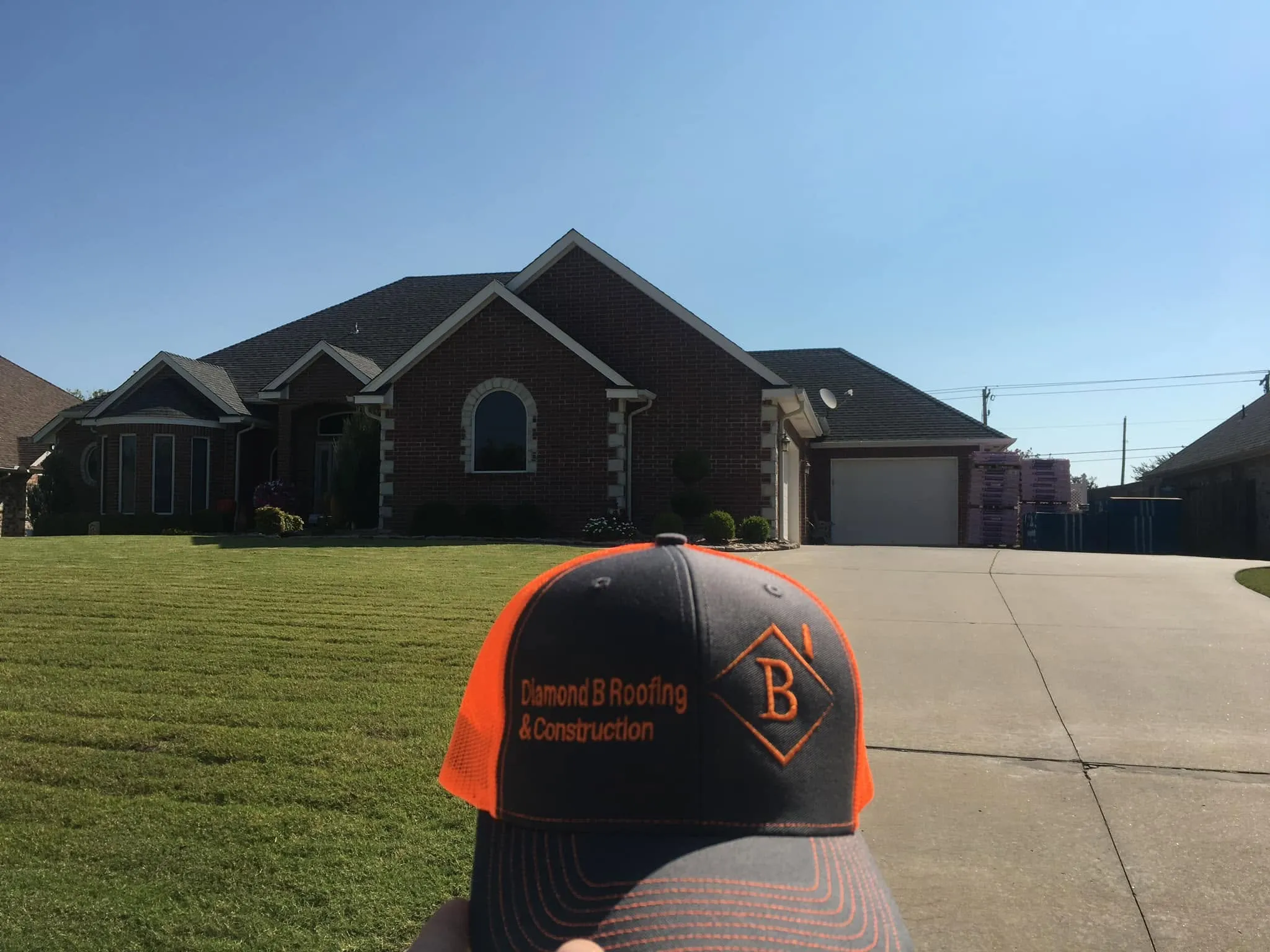 A Diamond B Roofing and Construction cap held up in front of a finished brick home